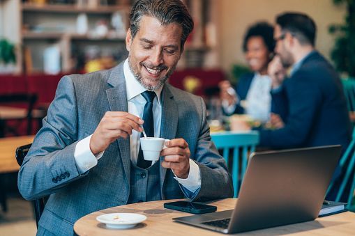 Lächelnder Mann in den Vierzigern in einem Café mit Laptop und Kaffeetasse, der eine ernsthafte Begegnung, einen positiven ersten Eindruck und authentische Kommunikation veranschaulicht.